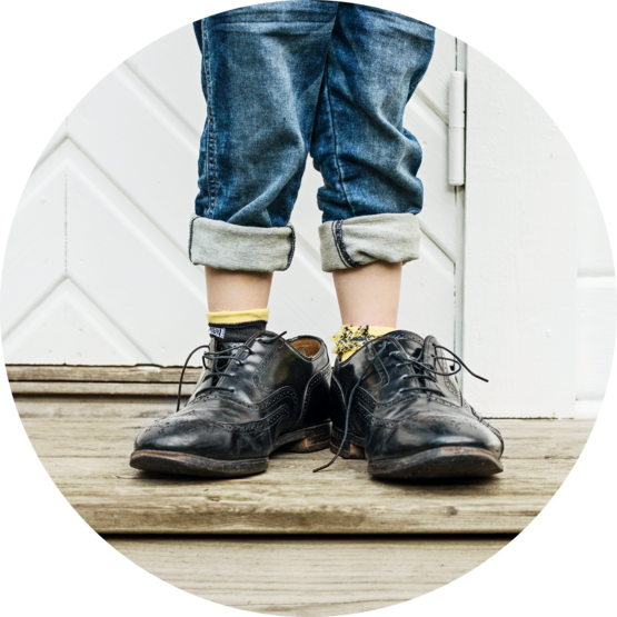 A close-up of a child's legs wearing rolled-up jeans and oversized black shoes, standing on wooden steps.