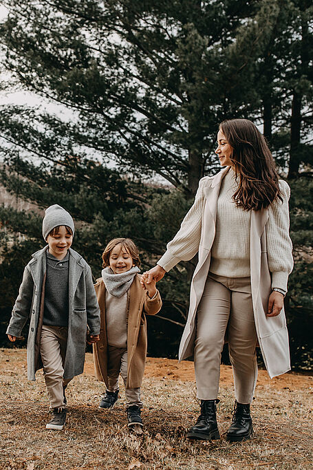 Mother and two children, all wearing coats and scarves, walking hand in hand through a forested area on a cloudy day.