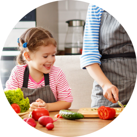 "A young girl in a striped shirt and apron smiling while watching an adult, also in an apron, chop vegetables in a kitchen. Fresh vegetables are spread out on the table.