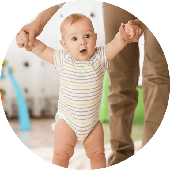 Baby in a striped onesie taking assisted steps while being held by an adult's hands, in a brightly lit room with toys in the background