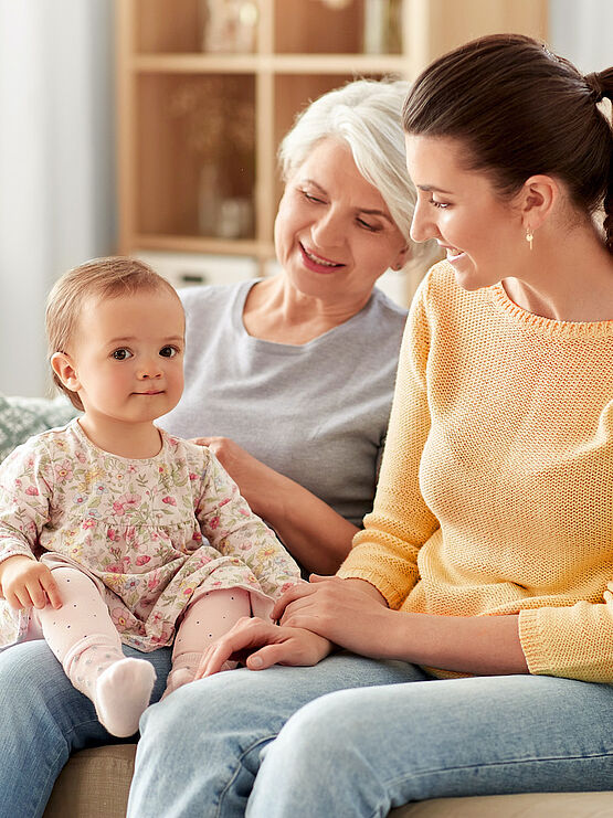 Three generations of women sitting on a couch: a grandmother, mother, and baby girl, enjoying each other's company at home