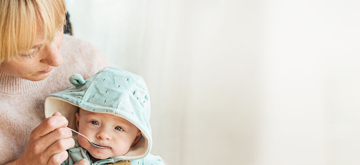 A baby in a light blue hoodie is being fed with a spoon by a woman.